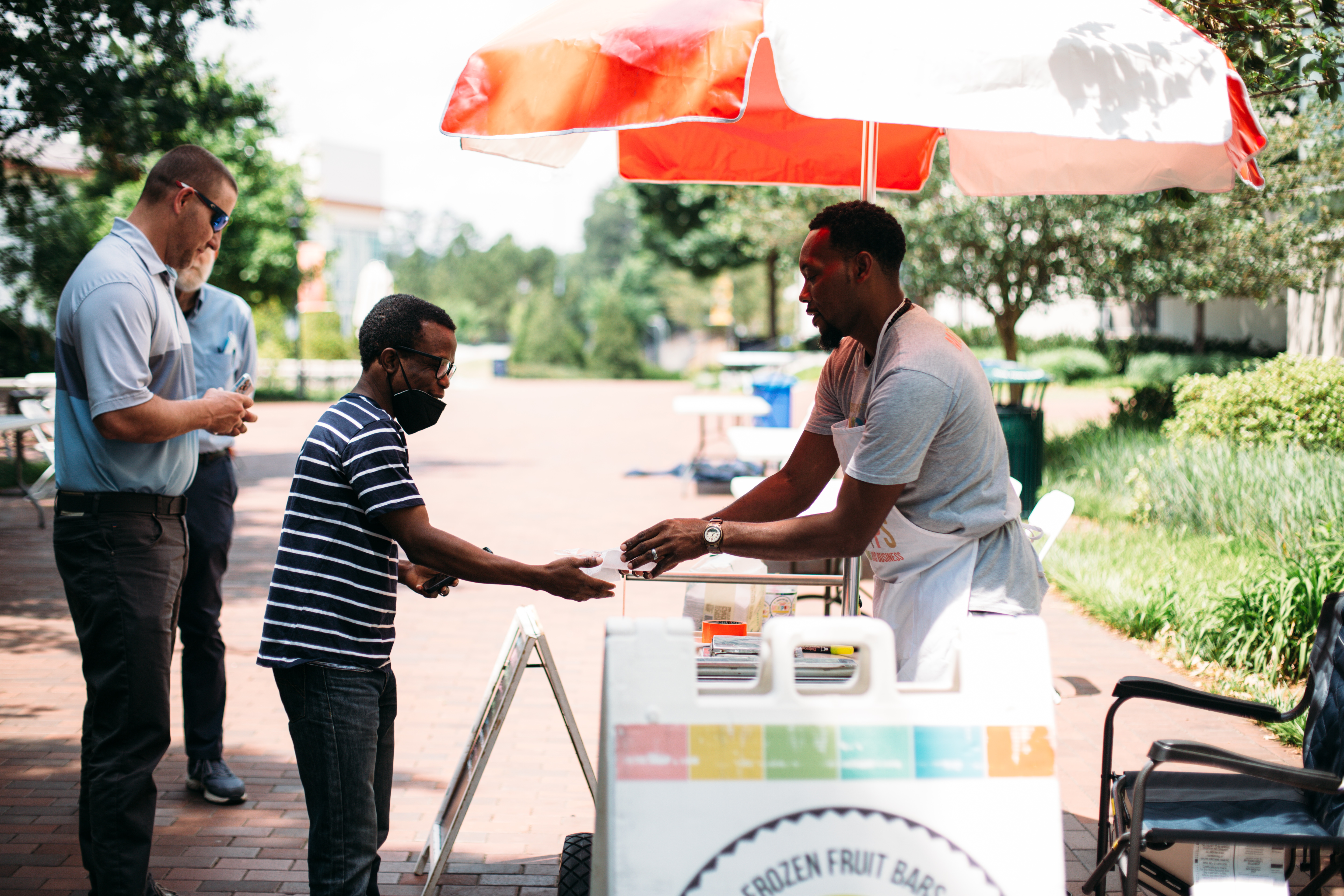 Man stands behind popsicle cart and hands another man a popsicle
