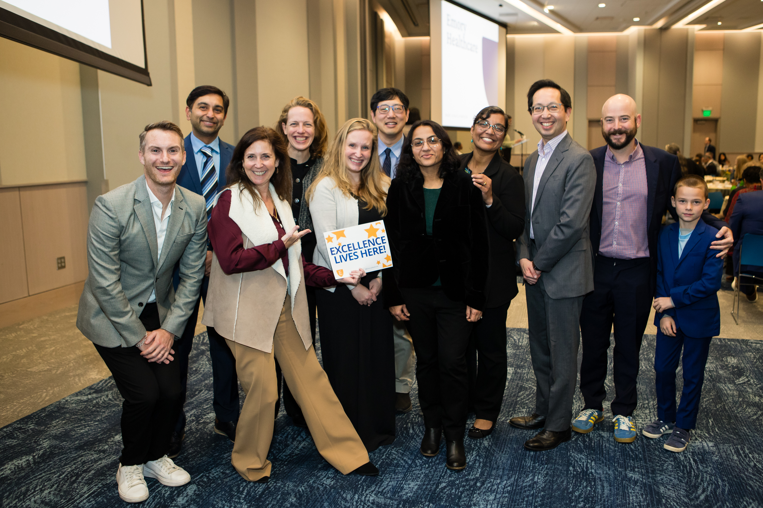 Large group of people smile for a photo with one woman holding up a sign that says "Excellence lives here!"