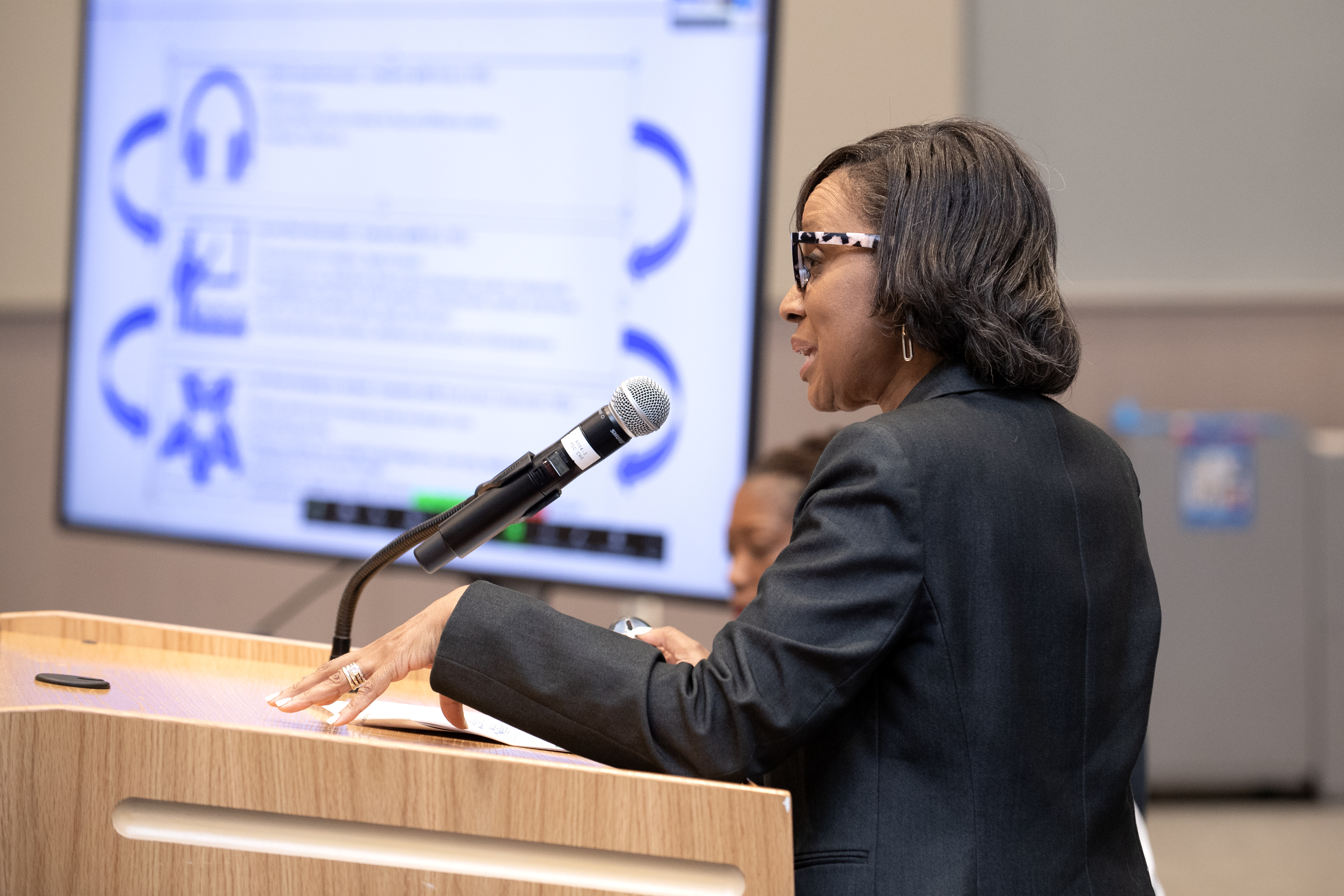 A woman presents with a PowerPoint screen next to her