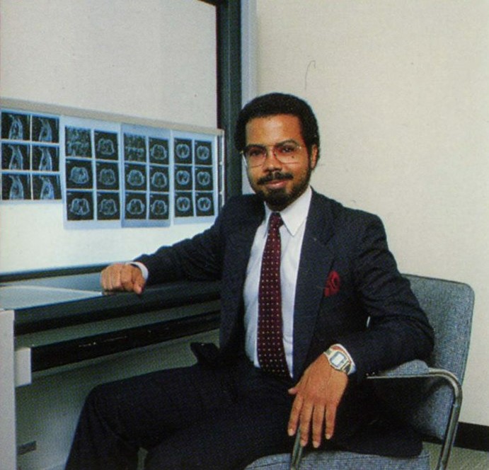 man with short dark hair moustache and beard and wearking square spectacles a dark suit jacket red tie and white shirt sitting beside a desk with images of a heart scan on a wall light box