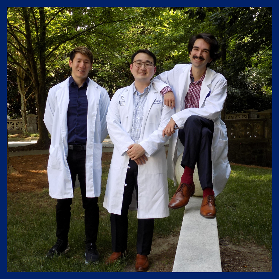 three young doctors wearing dark pants checked shirts of various colors and white coats over them standing in a shaded treeful area and smiling