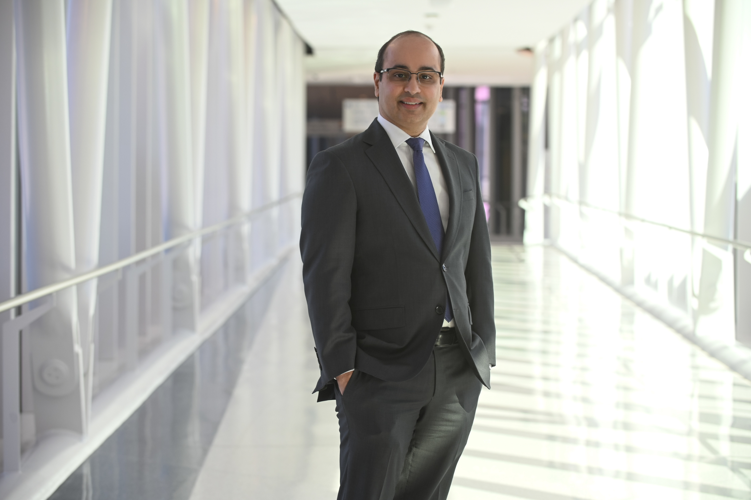 smiling man with short cropped hair and eyeglasses wearing a dark grey suit over a white shirt and blue tie standing in the middle of the covered pedestrian bridge made of white beams and glass