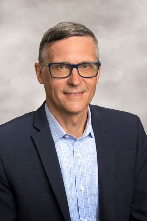 smiling person wearing spectacles and a navy sport coat over a light blue shirt