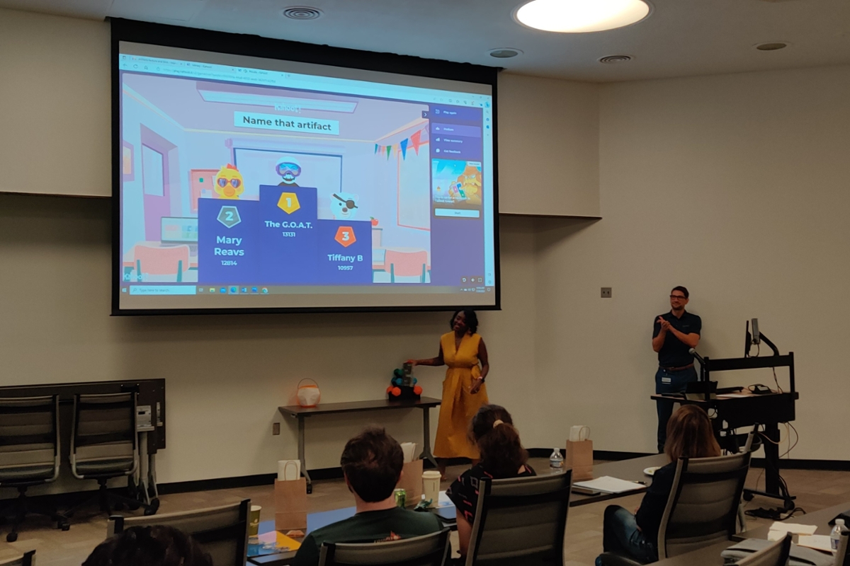 people sitting in darkened room listening to person in the front of the room and another person assisting with brightly colored slides projected on wall