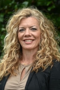 smiling person with long curly blond hair wearing a black jacket over a beige top and gold necklace
