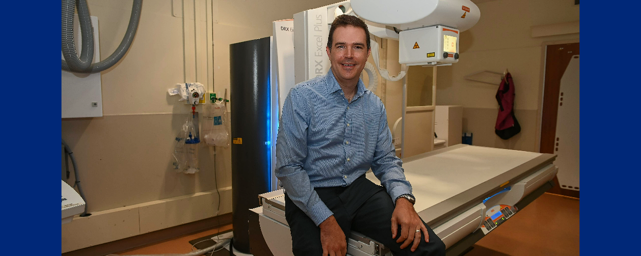 smiling person perched on the edge of an imaging table with the imaging machine behind them