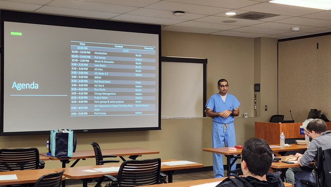 person standing at the front of a classroom wearing blue doctor scrubs and pointing to agenda on the board