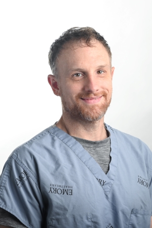 smiling man with reddish brown hair and facial hair wearing a blue scrubs top over a grey tshirt