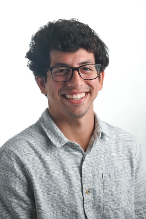 smiling man with short curly brown hair wearing dark rimmed glasses and a light grey checked shirt