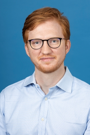 smiling man with short red hair wearing black rimmed round spectacles and a light blue shirt