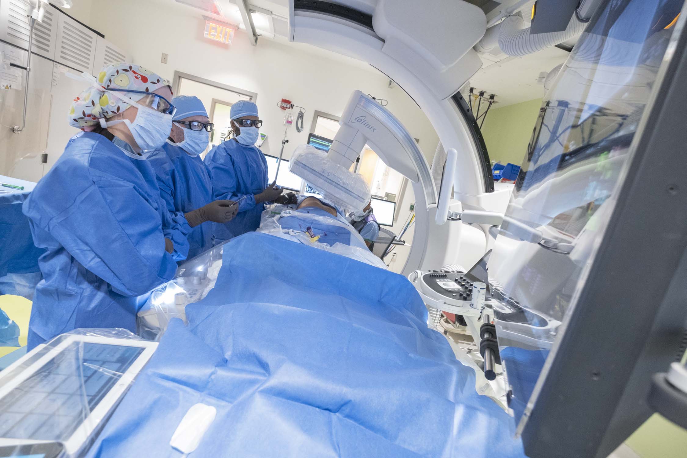 three radiologists wearing blue scrubs and caps and protective eyewear and masks covering their noses and  mouths performing an image-guided procedure