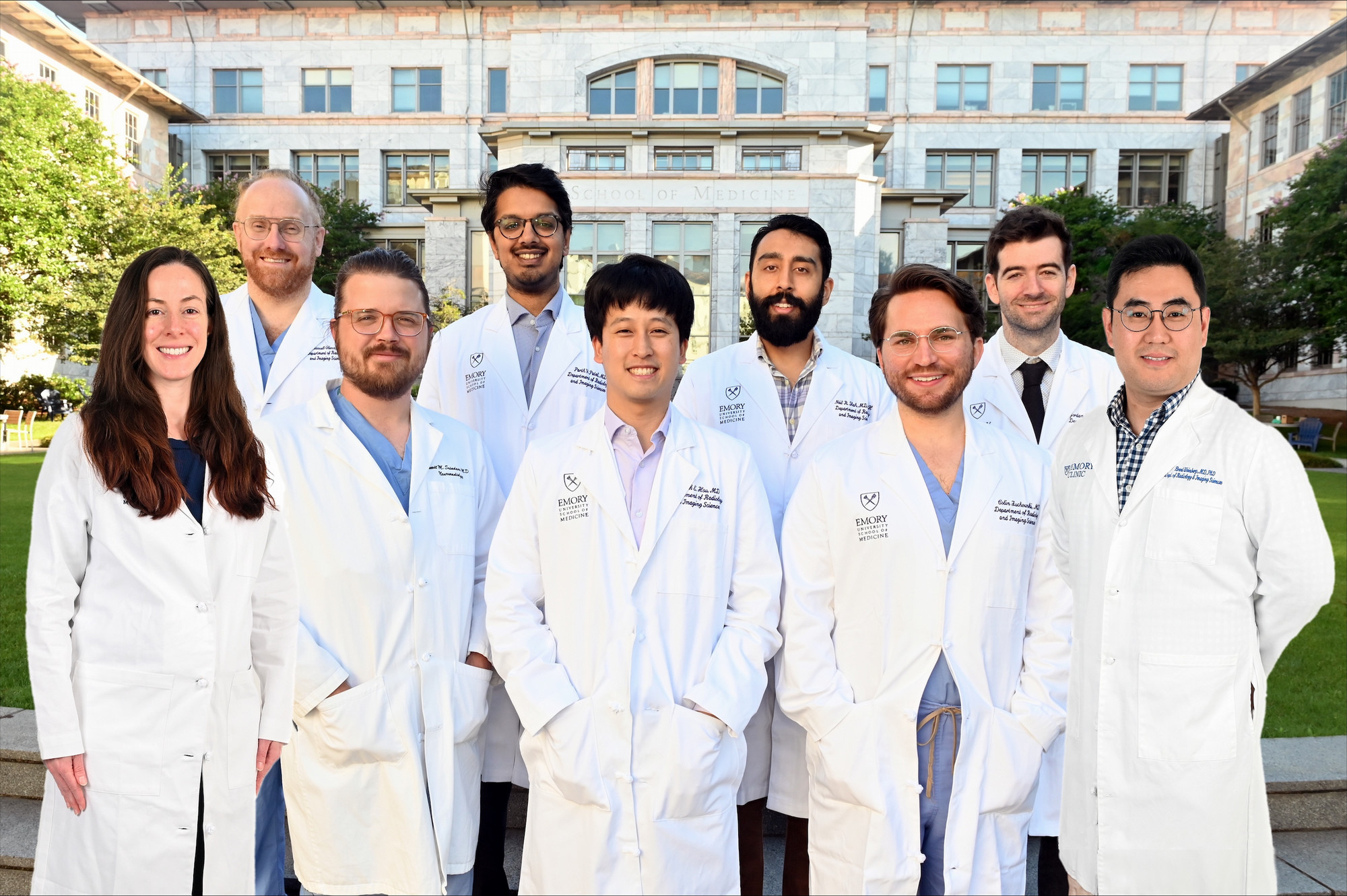 nine people standing in front of the glass and marble Emory Medicine building smiling and wearing white doctor coats