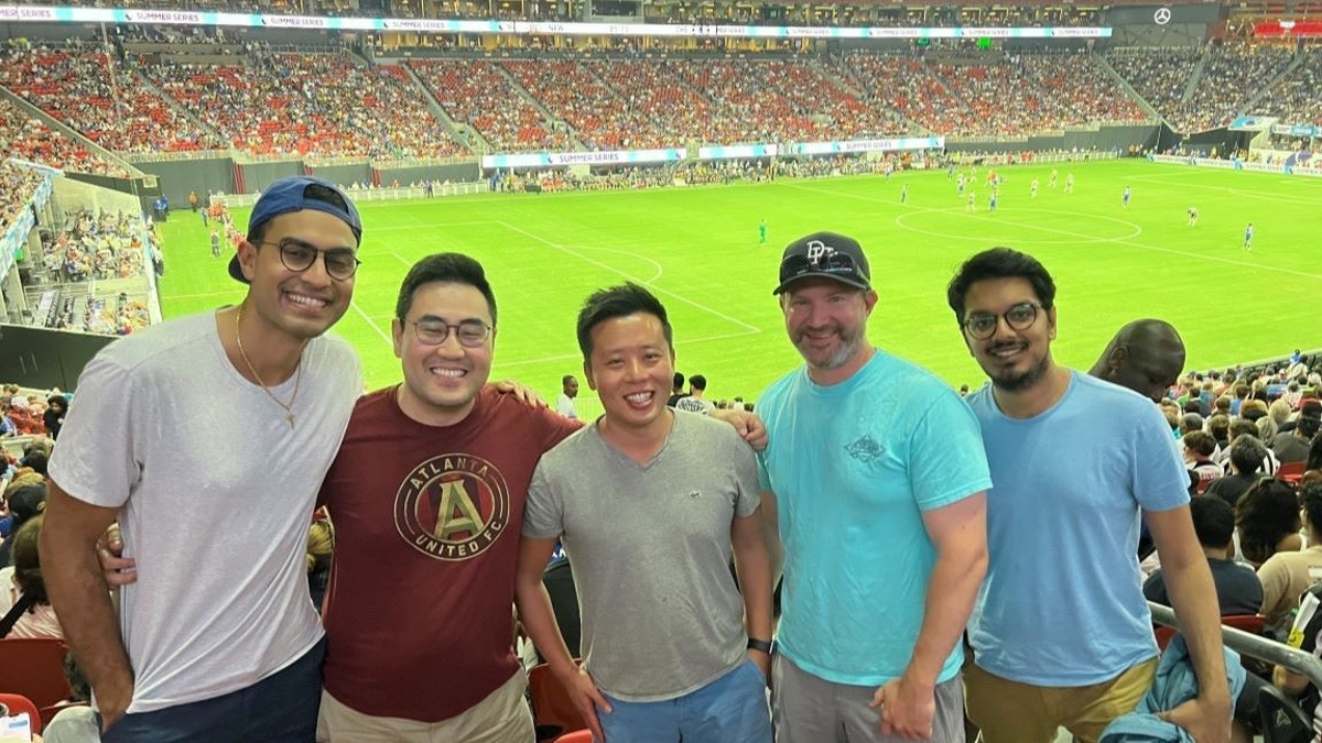 five people standing in a row smiling in the stands with the soccer pitch and the rest of the stadium crowd behind them