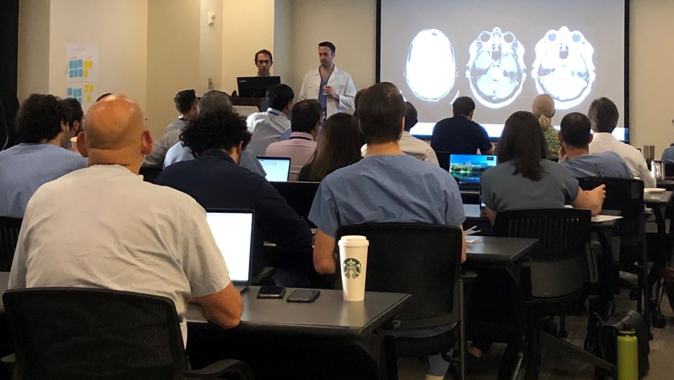 people seated in a classroom with two people at the front of the room with images of head scans projected on front wall