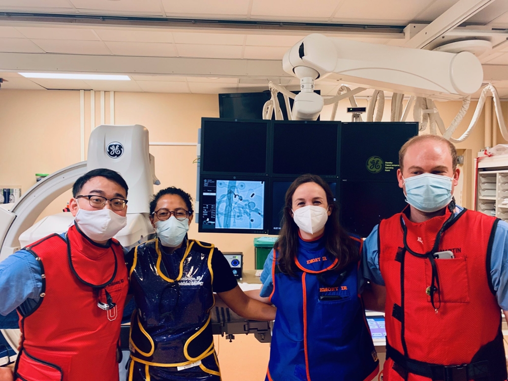 2 men and 2 women wearing bright red and blue aprons that protect them against radiation and masks standing with their arms linked in front of imaging equipment