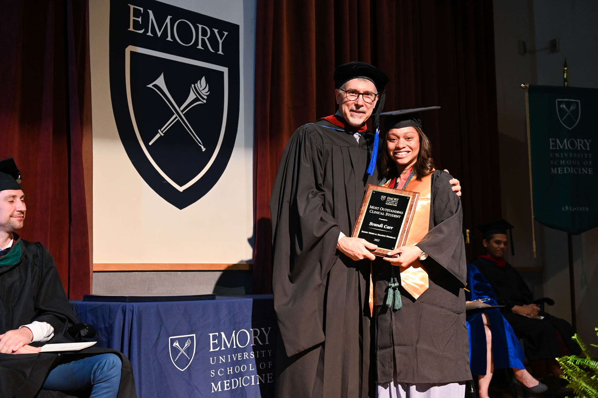 student in graduation cap and gown holding certificate beside instructor with Emory seal in the background