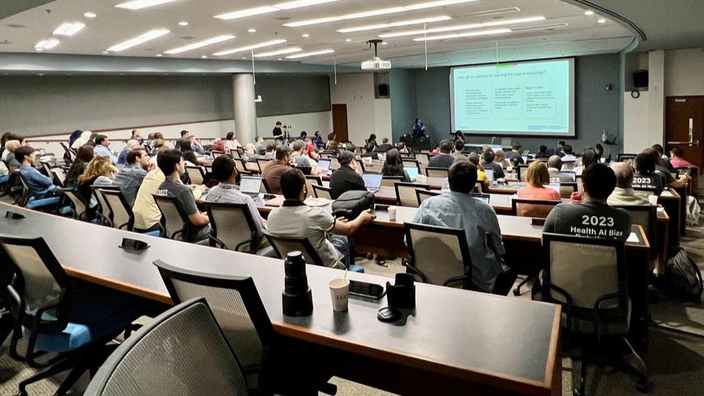 large lecture hall filled with people using laptops and viewing screen in the front of the room