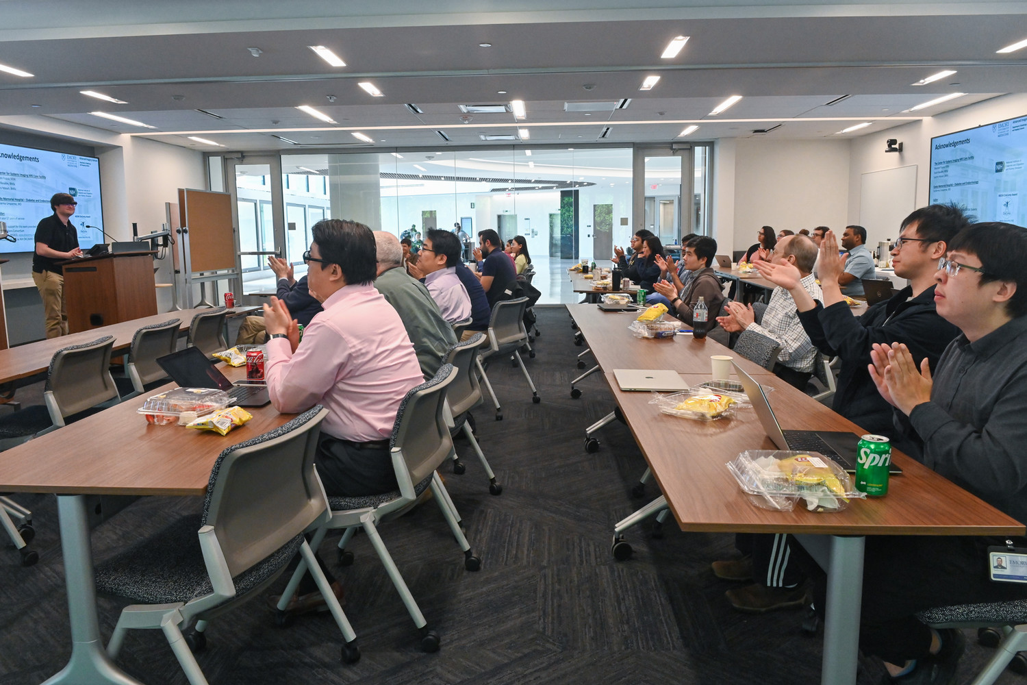 people sitting in two long rows of tables and chairs applauding person at the front of the room who is behind a lectern