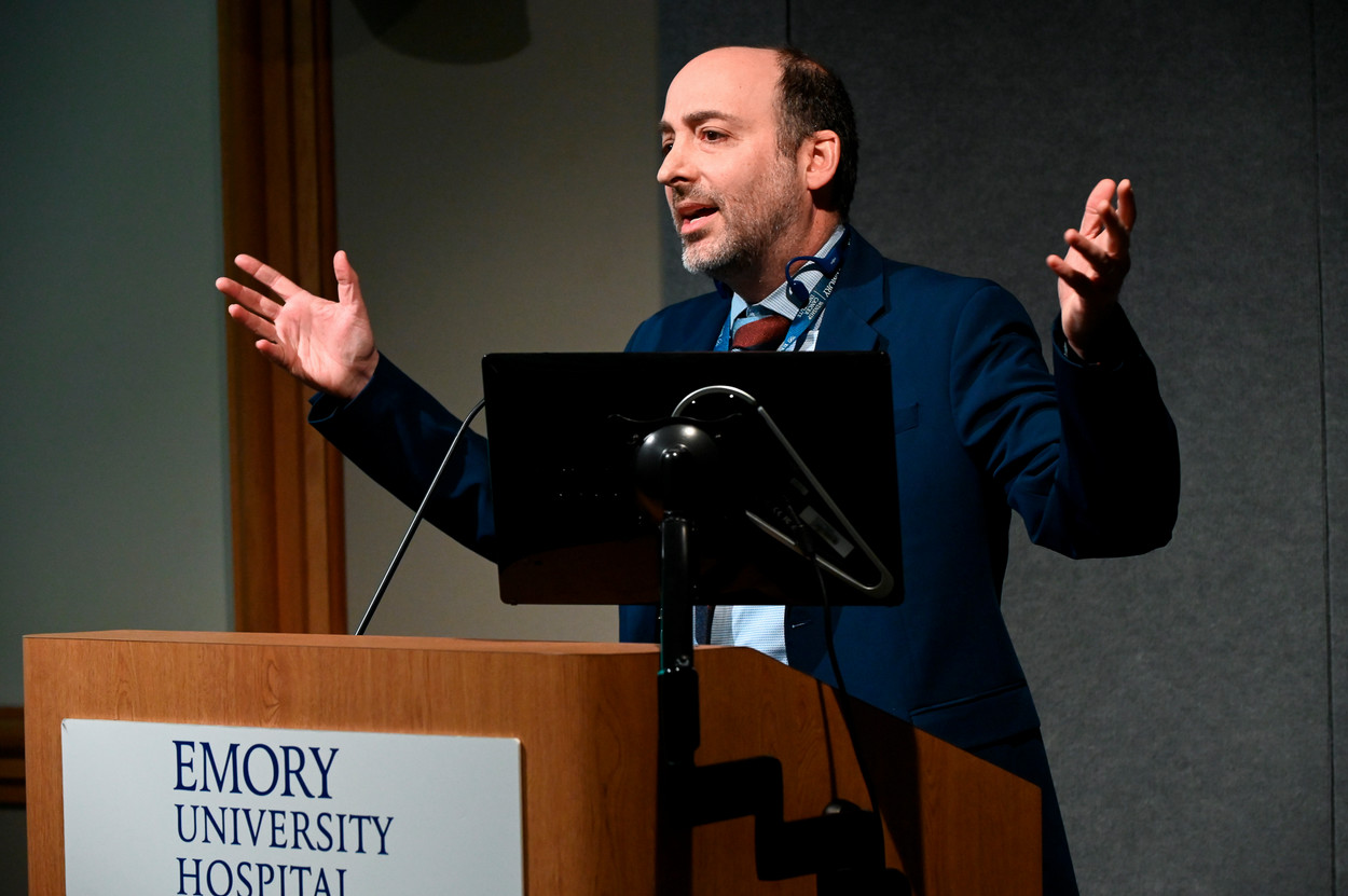 Person standing behind wooden lecturn with arms outstretched gesturing while they speak into a microphone attached to the lecturn