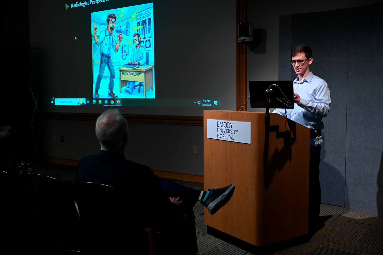 person talking at a wooden lecturn to an audience and there is a screen beside the speaker with a cartoon of an angry person yelling into a bullhorn