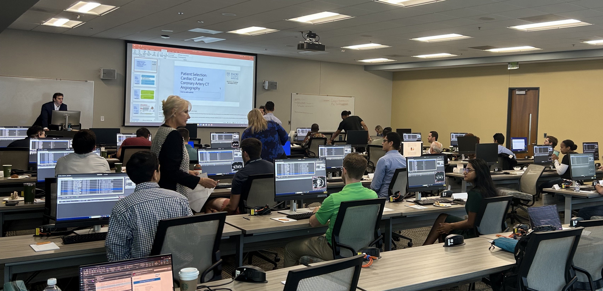 people in a classroom sitting and standing around computer workstations and talking with one another