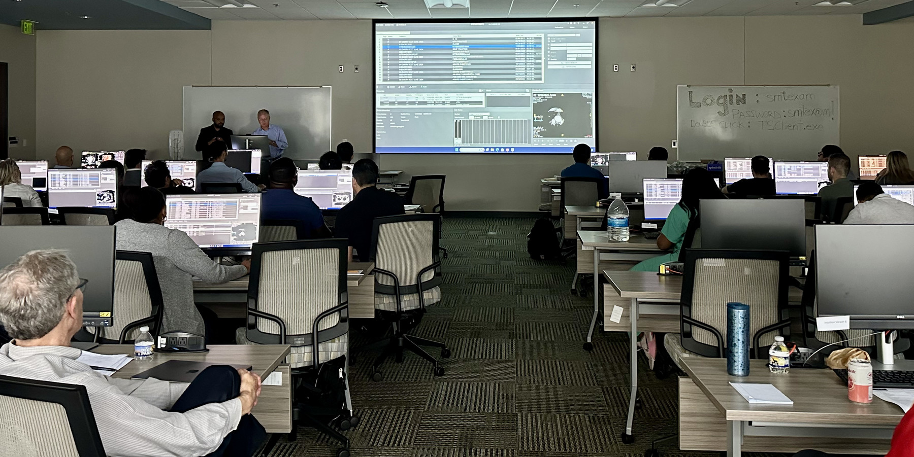people seated at workstations while two instructors talk at front of the room
