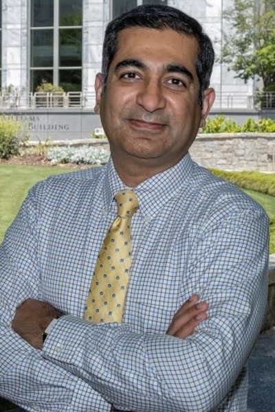 smiling man with short dark hair and arms crossed in front of his chest he is wearing a blue and white checked shirt and yellow tie