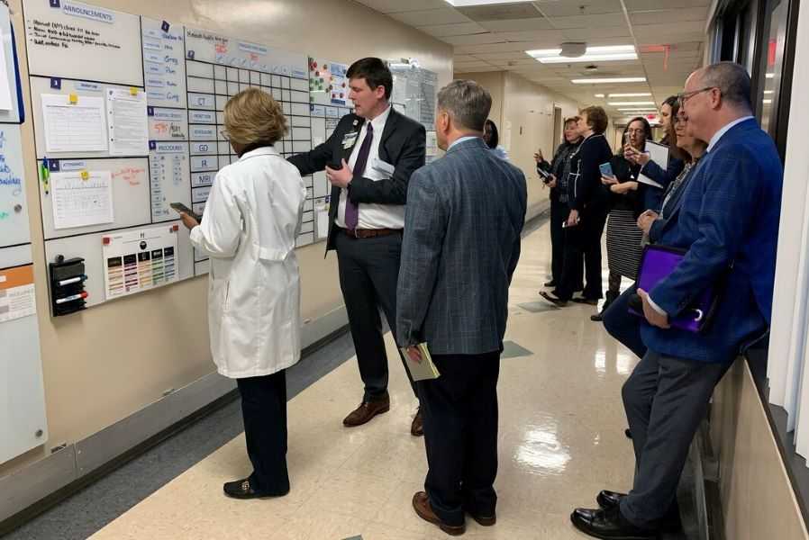 group of men and women standing in hospital hallway looking at a large board with words and charts on it to monitor workflow and people in that radiology work area