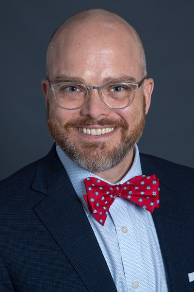 smiling man with bald head and blond and grey short beard and moustache wearing clear round glasses and a dark suit jacket over a medium blue shirt and red bowtie with silver dots