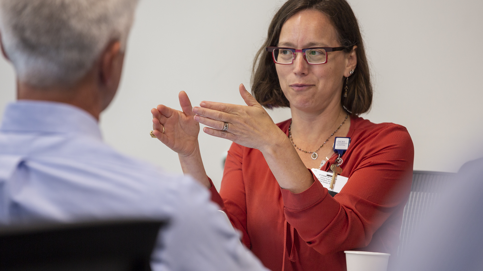 woman with shoulder length brown hair and glasses talking with her hands extended palms facing each other and suggesting distance between them while a man who we see only the back of his head listens