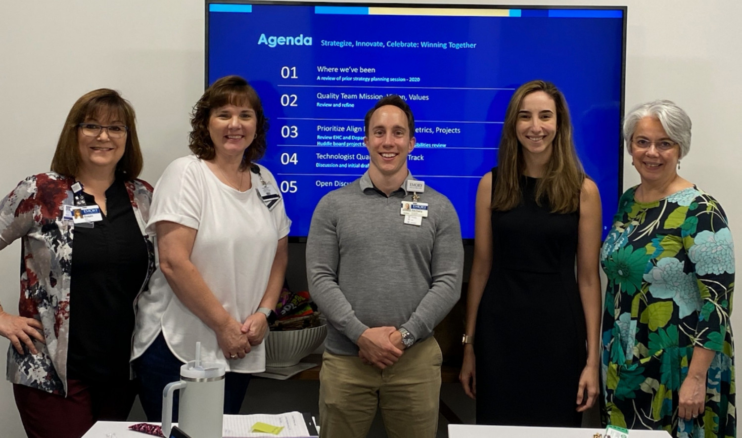 five people smiling and standing in front of a screen that has a schedule for a meeting on it