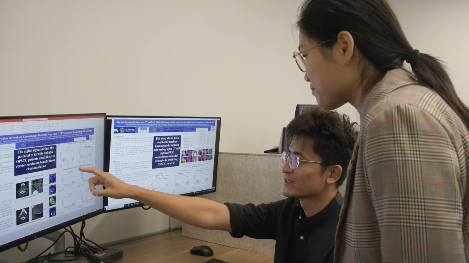 person seated in front of two computer monitors pointing at the monitors and talking to person standing beside them