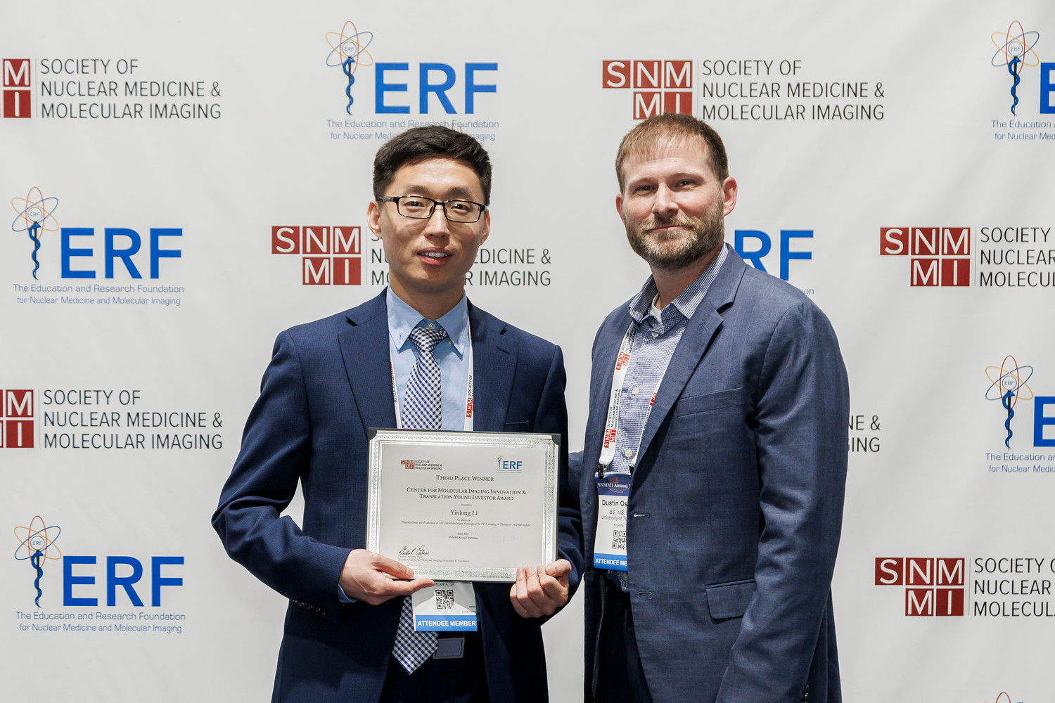 scientist holding award certificate standing beside another scientist in front of SNMMI banner