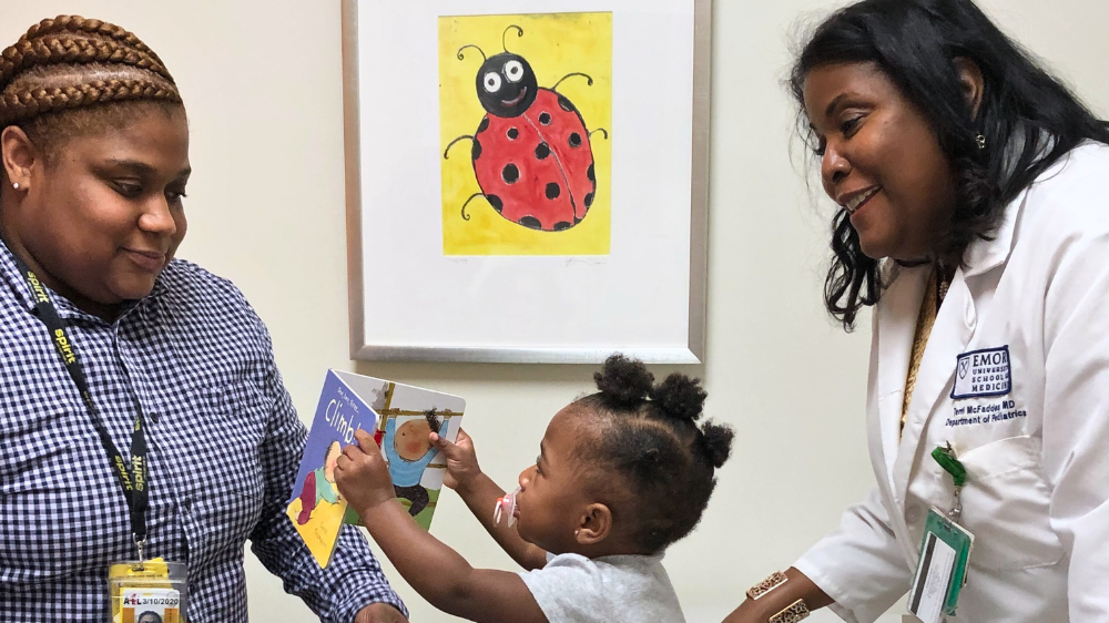 Two PARTNERS doctors are in a doctors office with a toddler, who is holding abook.
