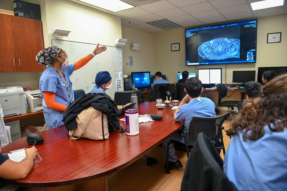 woman wearing hair covering and scrubs gesturing to large screen with radiology image while men and women sitting around conference table look at screen