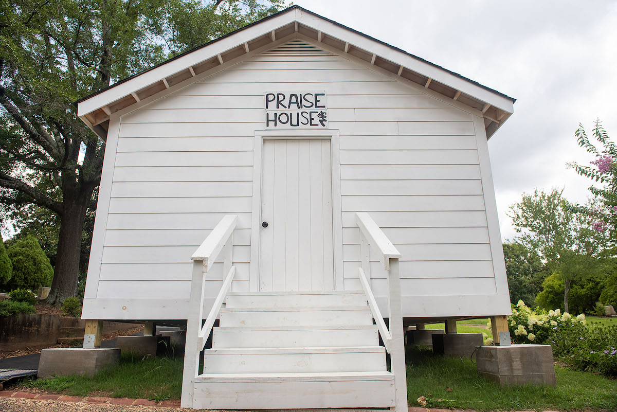 picture of white building in a grassy area with the words Praise House hand lettered on a sign over the door
