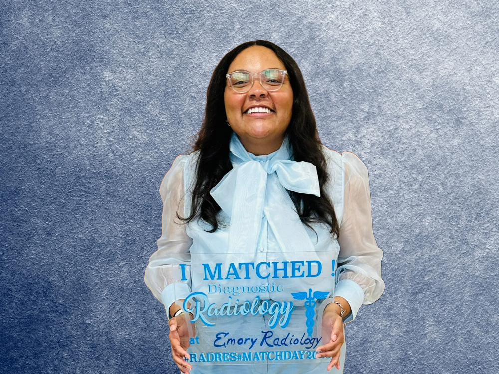 smiling person with long black hair wearing spectacles and a blue blouse with a large bow at the neckline holding a sign that says I matched diagnostic radiology Emory Radiology