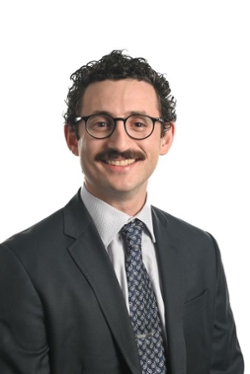 smiling person with short curly dark hair and moustache wearing round spectacles and dark suit jacket and tie over a light shirt