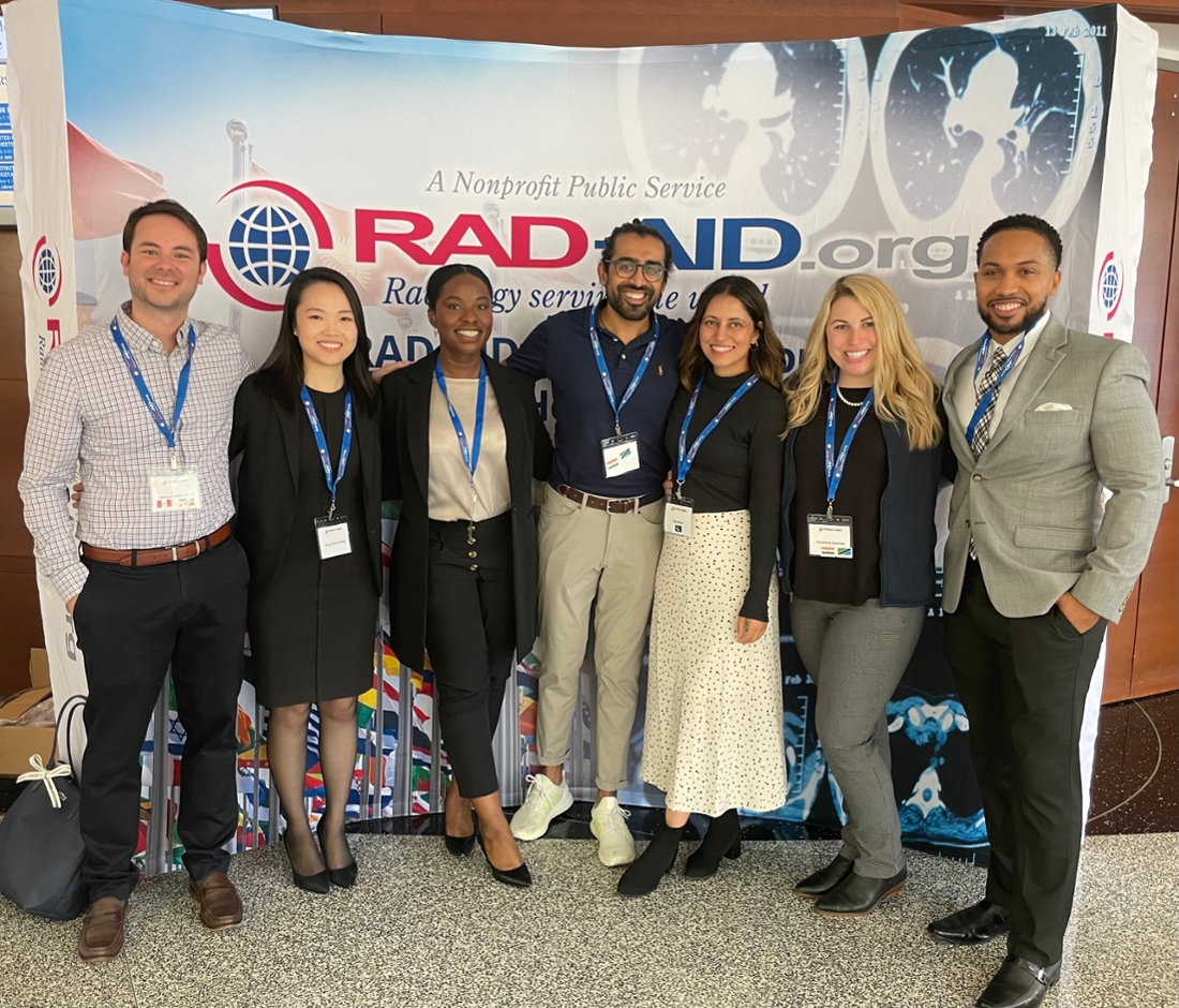 7 people standing in a row with their arms across each other's backs smiling at the camera with RAD-AID sign behind them