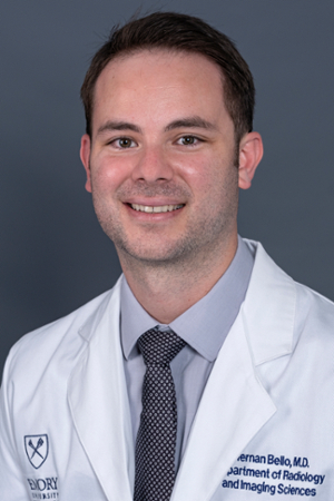 smiling man with facial stubble and short brown hair wearing a light grey shirt and black and grey tie under a white doctor coat that says Emory Healthcare and his name