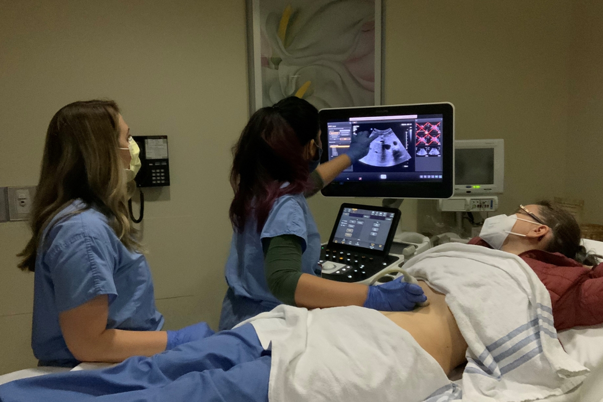 two women wearing blue doctor scrubs and masks look at an ultrasound screen while one holds a transducer to a female volunteers abdomen