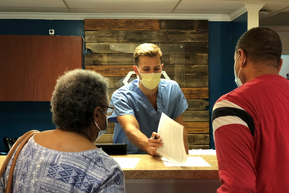 man in medium blue scrubs and wearing a face mask shows a woman and a man where to sign pices of paper at a reception desk in a clinic