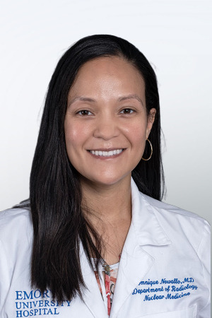 smiling woman with long dark hair down back and over her right shoulder wearing a white doctor coat over a floral top
