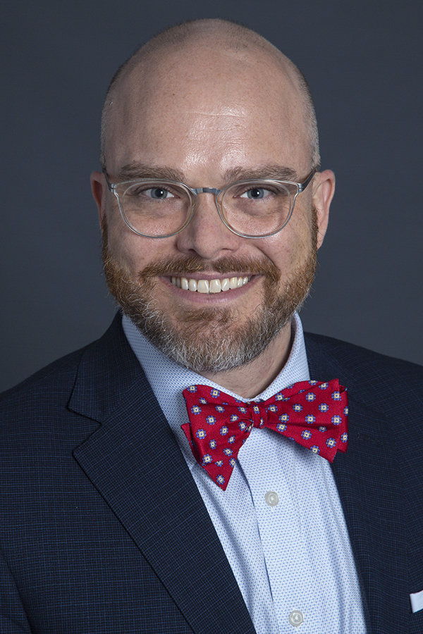 smiling man with bald head and reddish and grey beard and moustache wearing clear framed spectacles and a dark suit jacket over a light blue shirt and red and blue patterned bowie