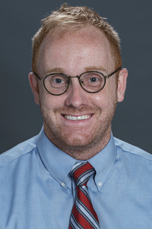smiling person wearing round spectacles and a french blue shirt with red and blue striped tie
