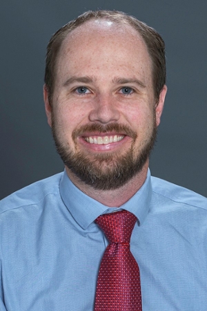 smiling person with light hair beard and moustache wearing a blue shirt and red tie