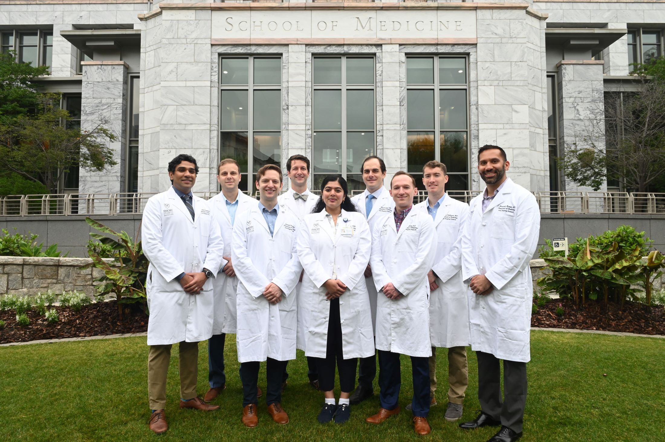 people wearing white doctor coats over pants and dress shirts standing in front of marble building with School of Medicine etched into the stone