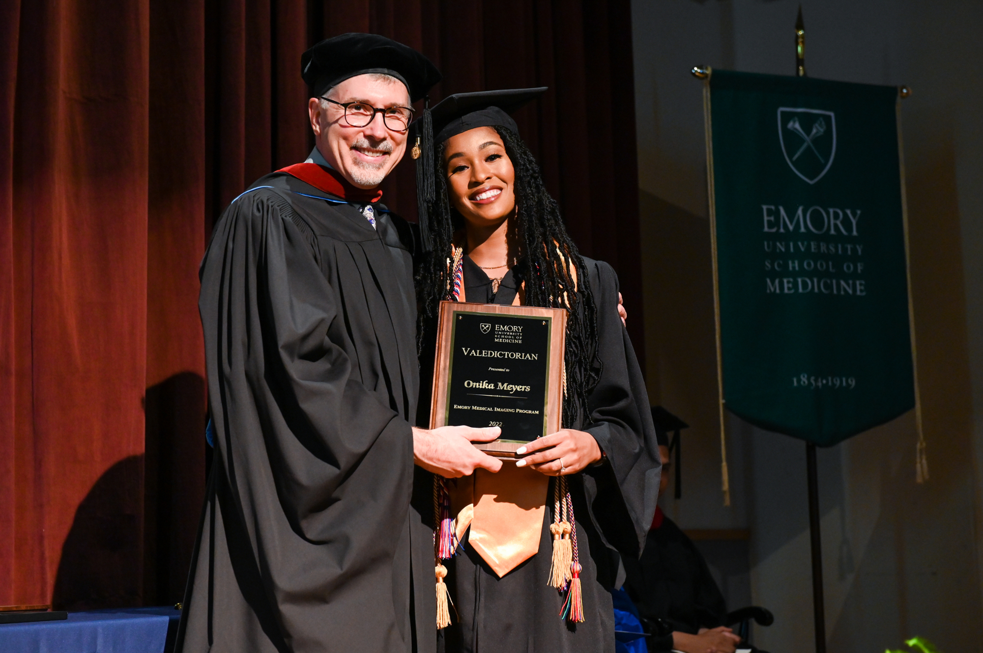 professor standing with student in graduation regalia holding valedictorian plaque