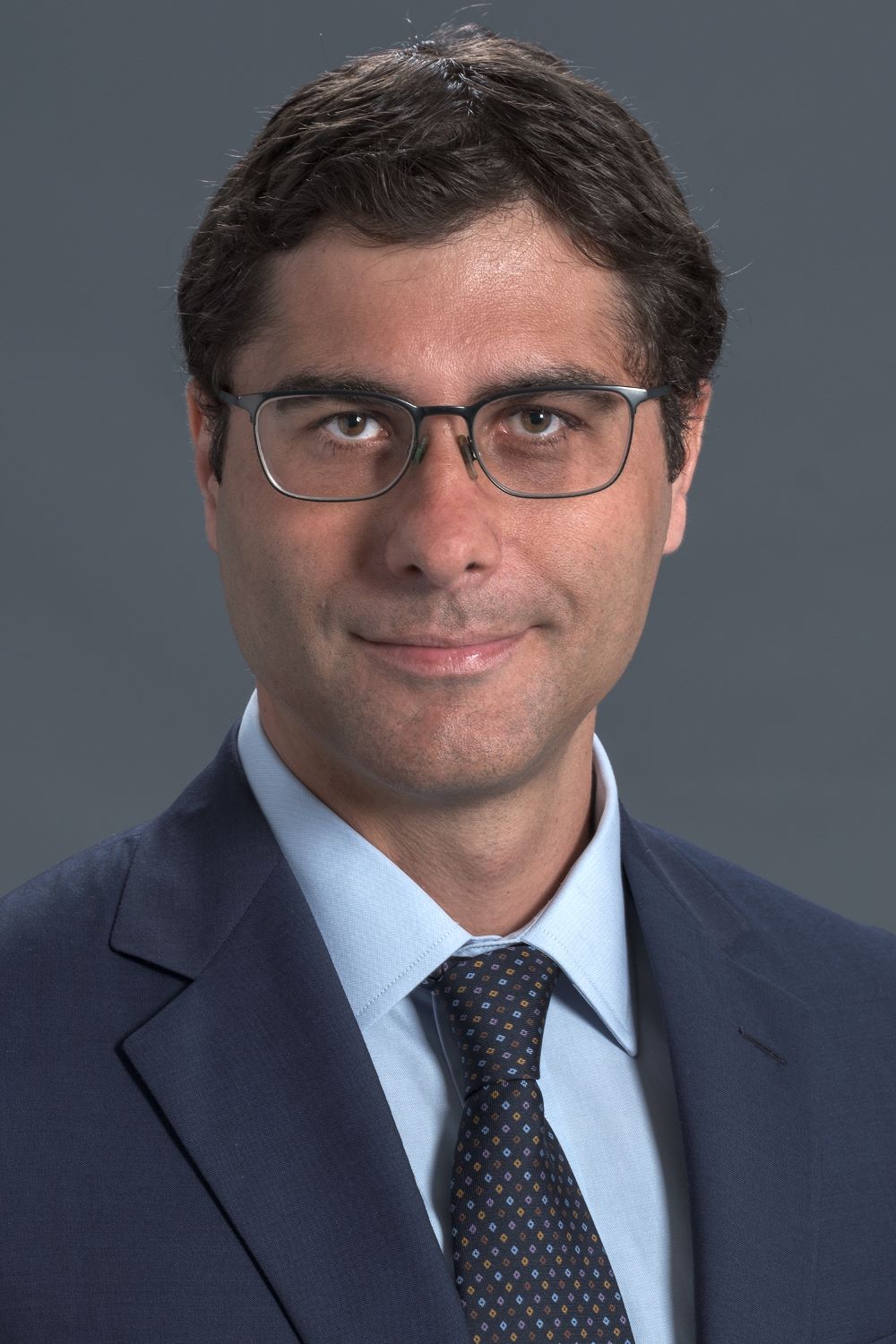 man with short dark hair and glasses wearing dark blue suit and tie with light blue shirt