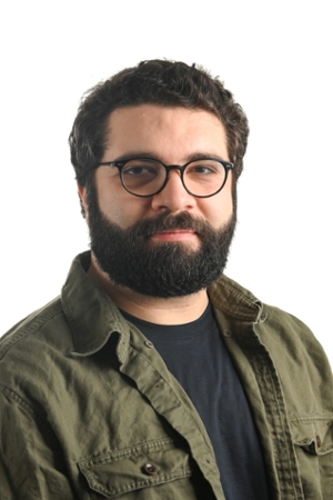 serious person with dark hair moustache and full beard wearing spectacles and an olive green jacket over a black shirt